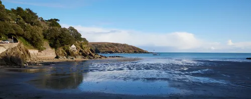 The image shows a landscape photograph taken from Bay 3 overlooking a quiet coastal beach in Devon, England, United Kingdom. The view was captured in the late afternoon during the winter season, as indicated by the low angle of the sunlight and the bare vegetation on the distant hillside. The beach is bordered by rocky cliffs and a stretch of sand with shallow pools of water reflecting the sky. The coastline, lined with trees and a stone wall, curves gently towards a headland projecting into the sea. The image highlights the natural beauty of the Devon coast, with clear skies and calm waters visible in the distance.
